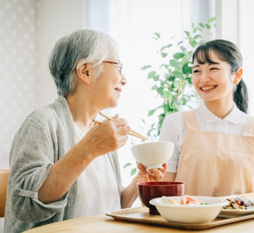 食卓で食事をとる高齢の女性と、隣で声をかける職員