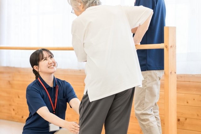 平行棒につかまって歩く練習をする高齢の女性と、笑顔で見守る職員