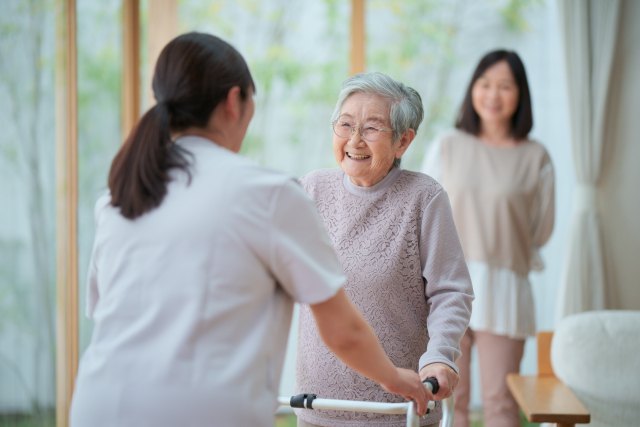 歩行器で歩く高齢の女性と、そばで支える職員